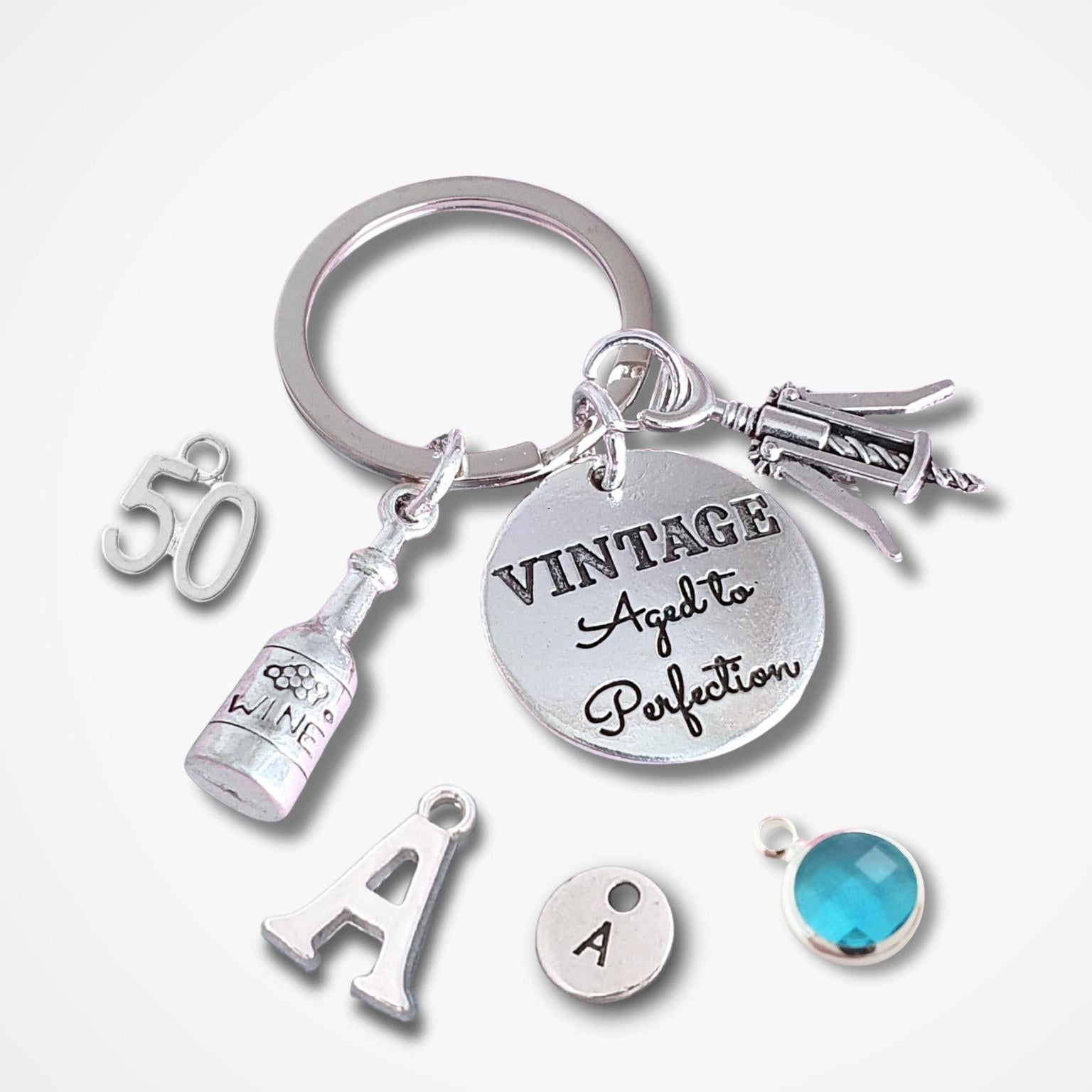 A silver-tone keychain with multiple charms, including a round disc engraved 'VINTAGE Aged To Perfection,' a bottle charm, a '50' charm, an 'A' letter charm, a small round 'A' charm, and a faceted blue gemstone charm, shown on a light gray background.