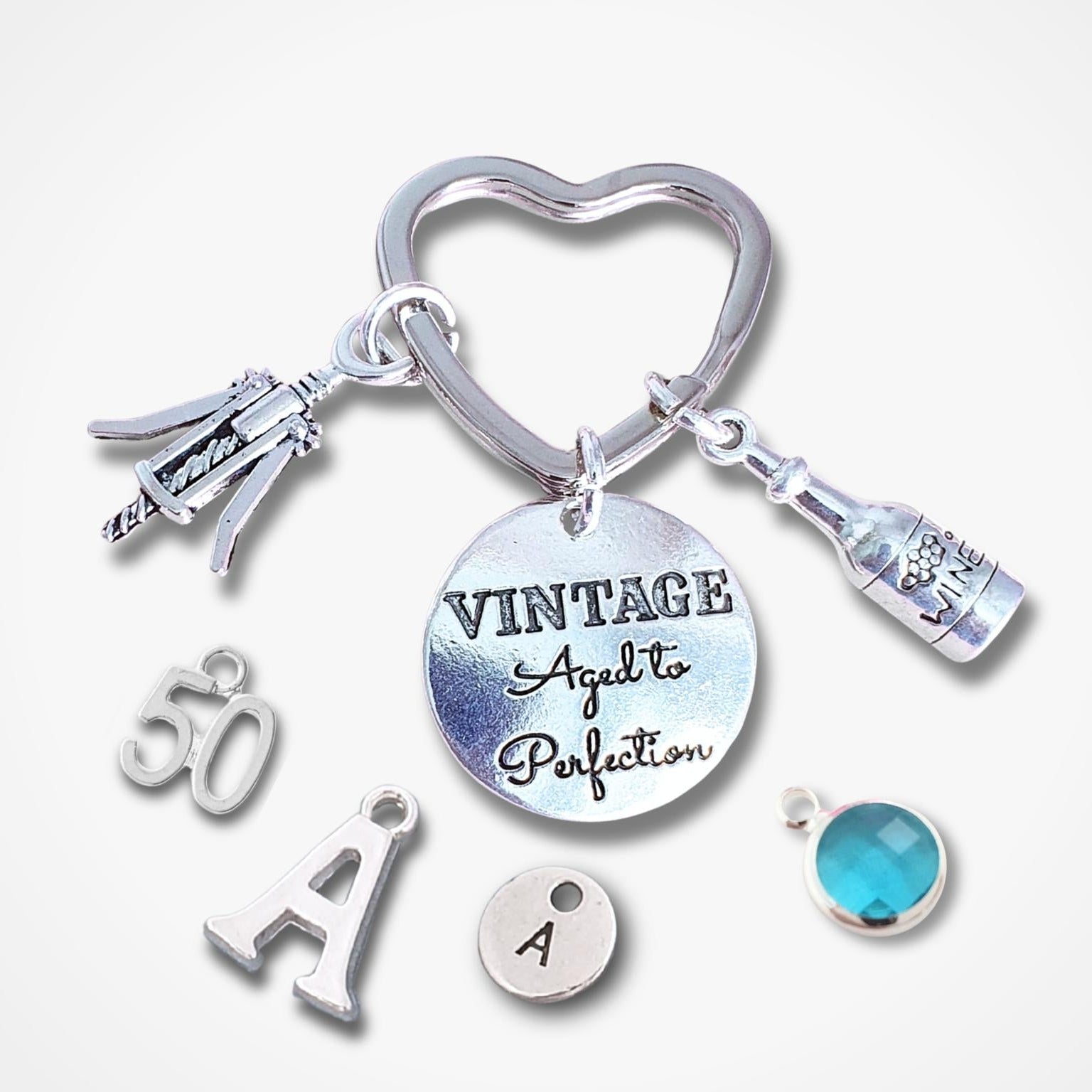 A silver charm keychain with multiple personalized and decorative charms, including a heart-shaped ring, a round charm engraved with 'VINTAGE Aged to Perfection', a bottle charm, a corkscrew charm, a '50' charm, an 'A' charm, a small round charm with the letter 'A', and a turquoise blue faceted bead.