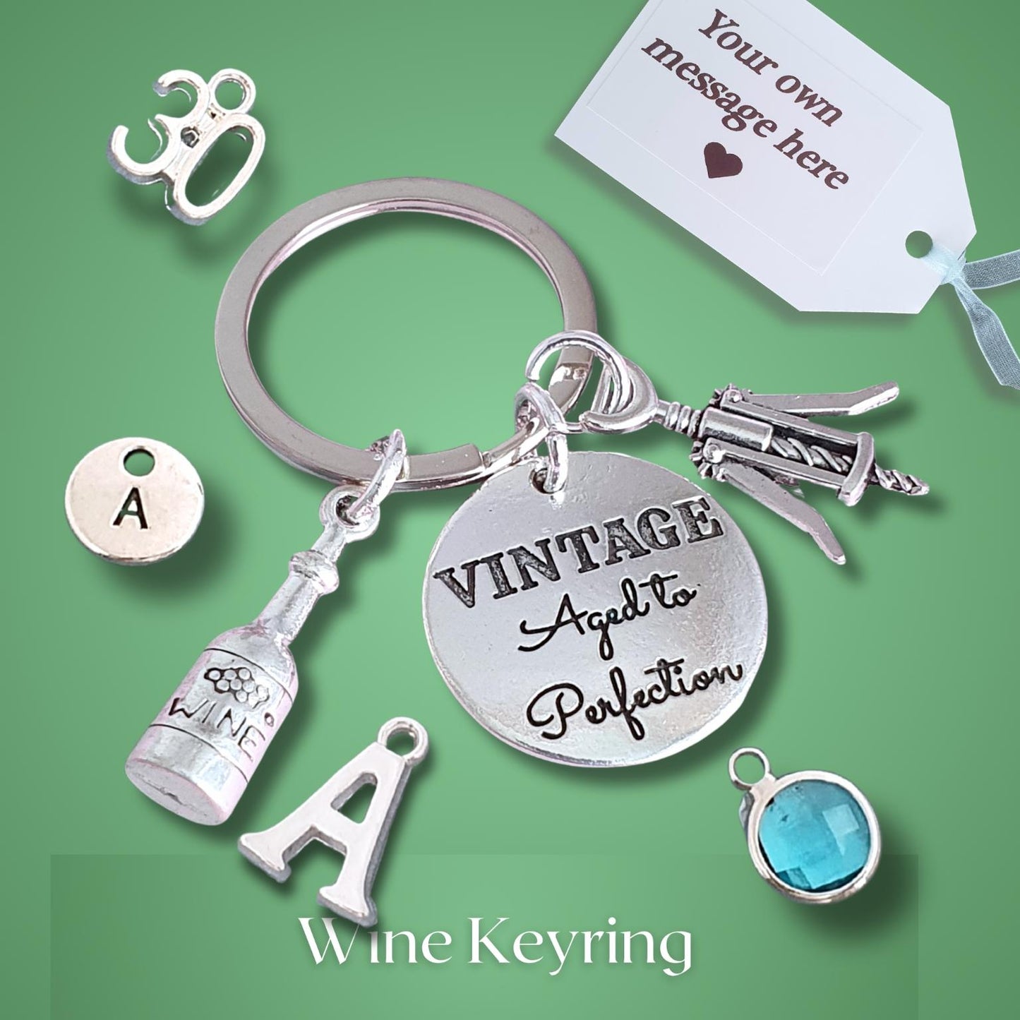 A silver-tone wine-themed keyring with multiple charms, including a round engraved disc reading 'VINTAGE Aged to Perfection,' a corkscrew, a wine bottle charm, two letter 'A' charms, and a small blue gemstone charm, displayed on a green background with a white message tag.