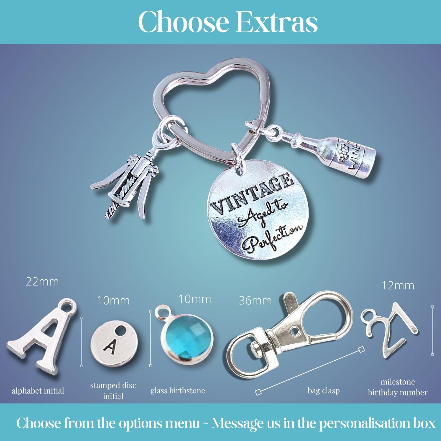 Silver keychain customization options under the heading 'Choose Extras' on a blue gradient background. The main product shown is a silver heart-shaped key ring with charms including a script nameplate reading 'VINTAGE Aged to perfection,' a bottle charm, and corkscrew.