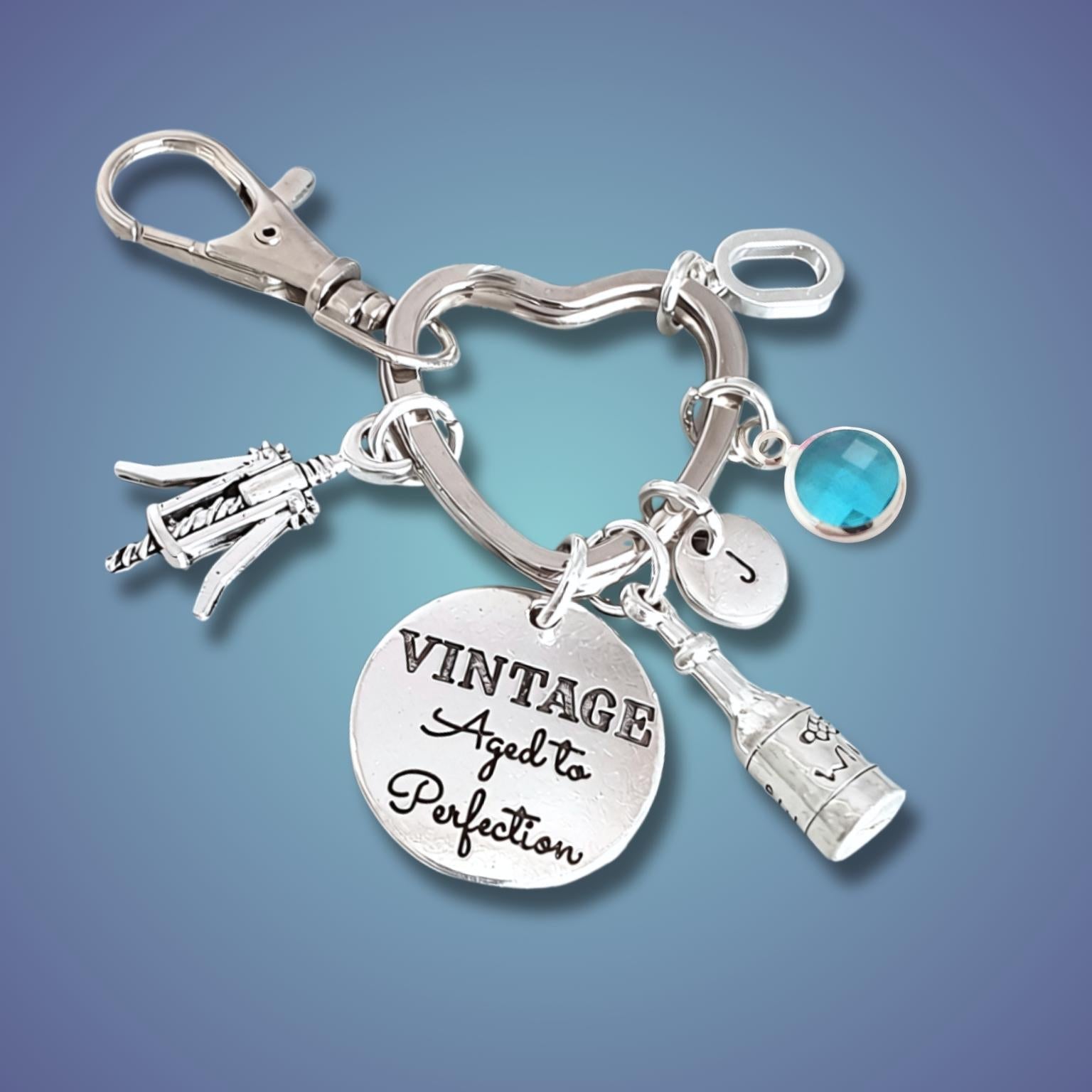 A silver charm keychain featuring a lobster clasp, a heart-shaped ring, a round engraved disc reading 'VINTAGE Aged to Perfection', a bottle charm, a small silver charm, and a round blue gemstone accent, shown on a blue gradient background.