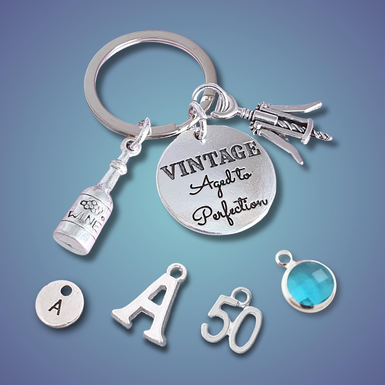 A silver-tone keychain charm set displayed on a blue gradient background. The main keychain features a round engraved disc reading 'VINTAGE Aged to Perfection,' a bottle-shaped charm, a corkscrew charm, and a key ring.