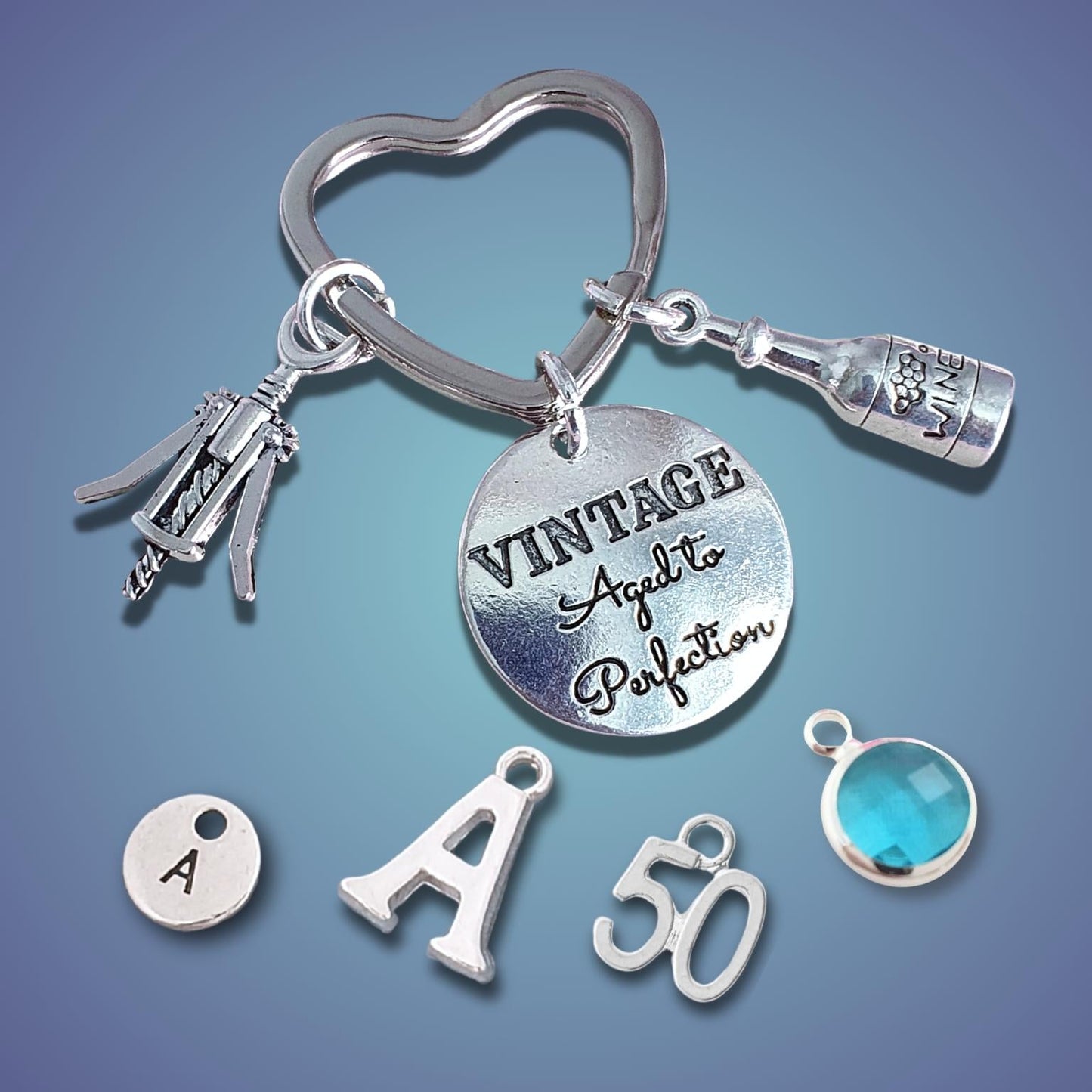 A silver-tone keychain set with charms, including a heart-shaped ring, a bottle charm, a corkscrew charm, a round engraved disc reading 'VINTAGE Aged to Perfection,' a small round charm marked 'A,' a letter 'A' charm, a '50' charm, and a faceted blue gemstone charm, displayed on a blue gradient background.