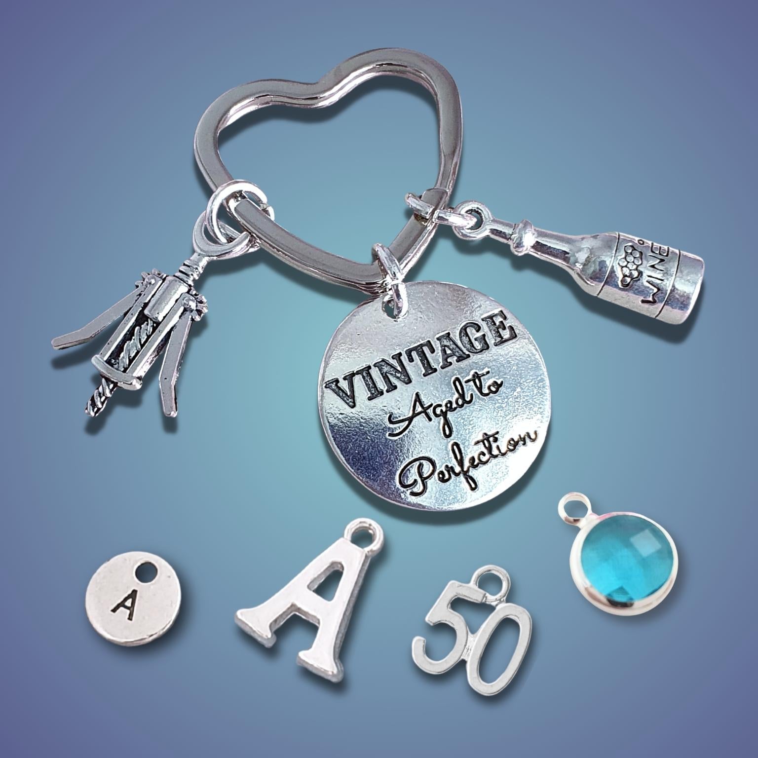A silver-tone keychain set with charms, including a heart-shaped ring, a bottle charm, a corkscrew charm, a round engraved disc reading 'VINTAGE Aged to Perfection,' a small round charm marked 'A,' a letter 'A' charm, a '50' charm, and a faceted blue gemstone charm, displayed on a blue gradient background.