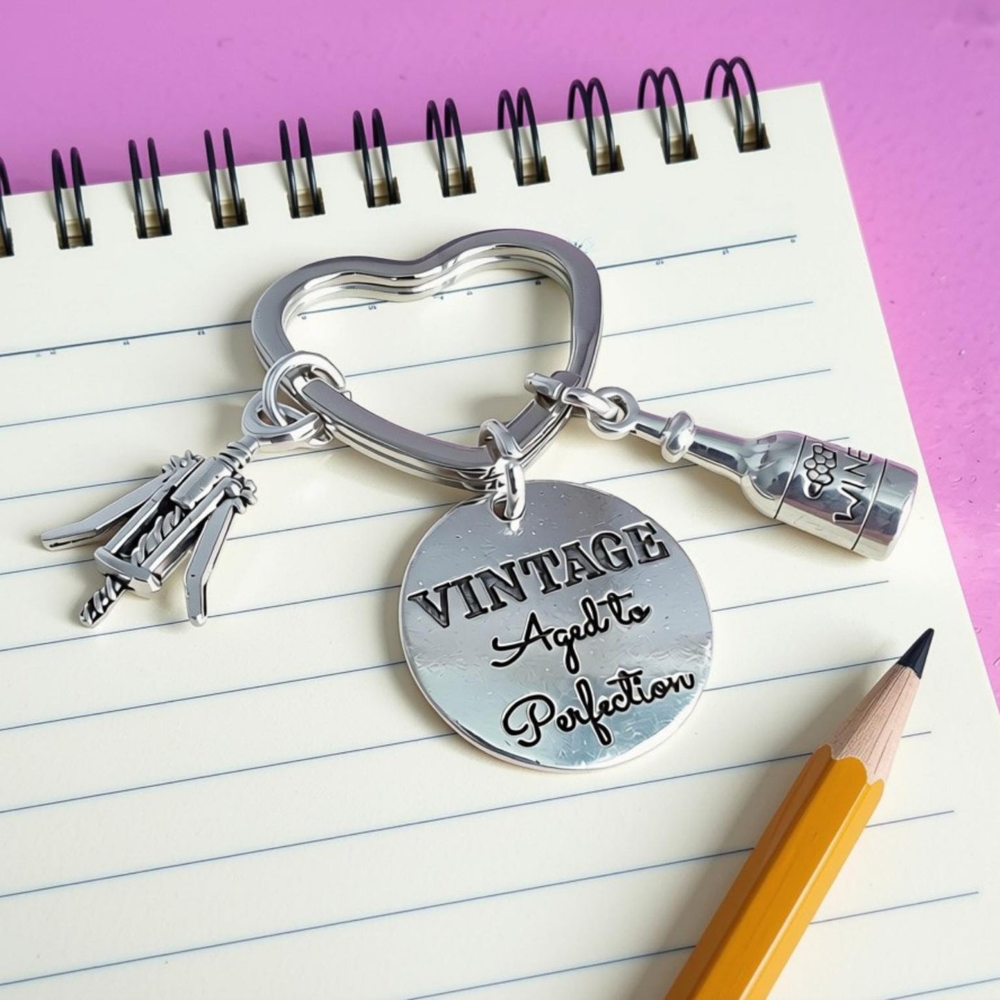 A silver metal keychain with charms, including a heart-shaped ring, a bottle-shaped charm, and a round disc engraved with 'VINTAGE Agreed Perfection', displayed on an open lined notebook with a yellow pencil on a pink background.