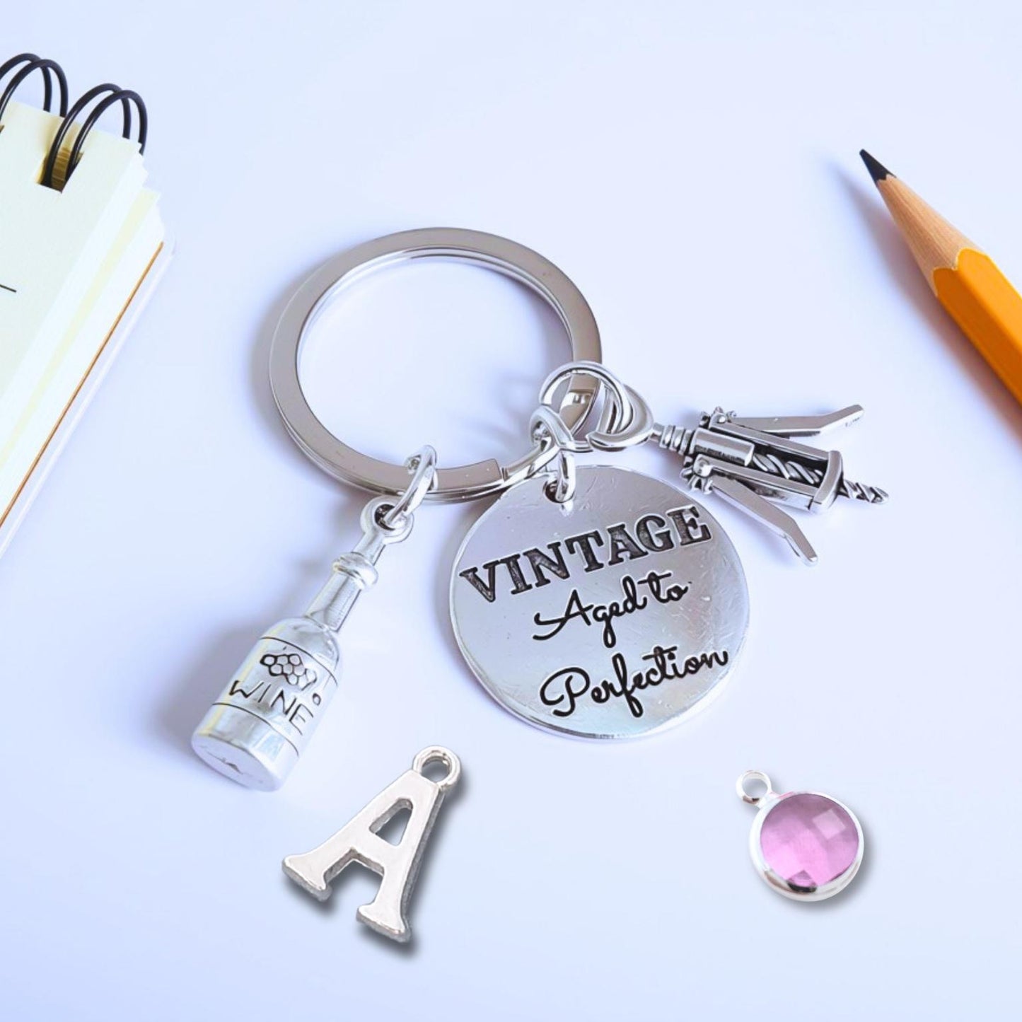 A silver metal keychain with charms, including a round engraved disc that reads 'VINTAGE Agreed to Perfection,' a bottle charm, a keyring, and a small pink gem charm, styled on a white surface with a pencil, notebook, and letter 'A' charm nearby.