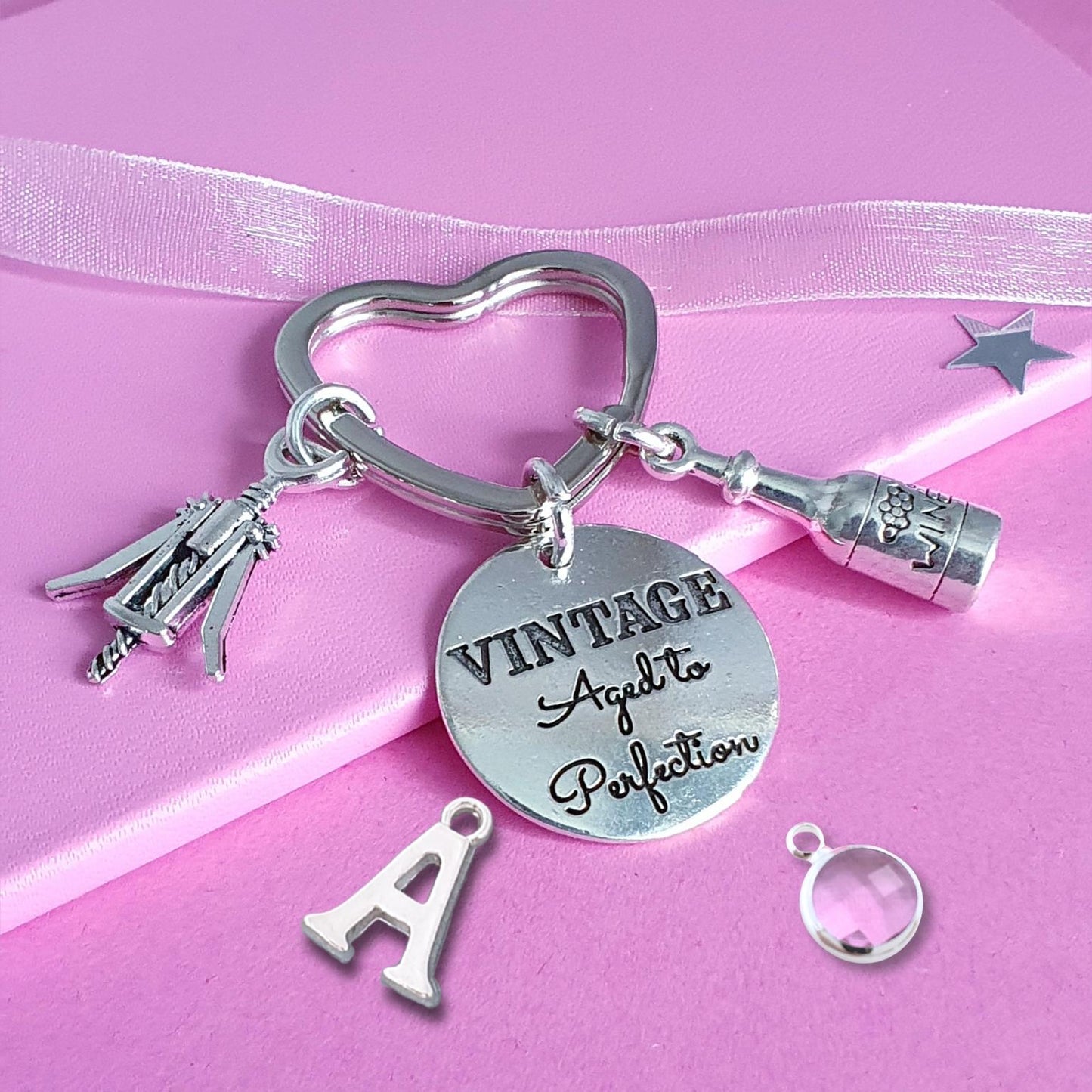 A silver charm keychain set displayed on a pink ribbon. The keychain features a heart-shaped ring, a round engraved disc reading 'VINTAGE Aged to Perfection', a bottle charm, a small corkscrew charm, a keyring, and a letter 'A' charm.