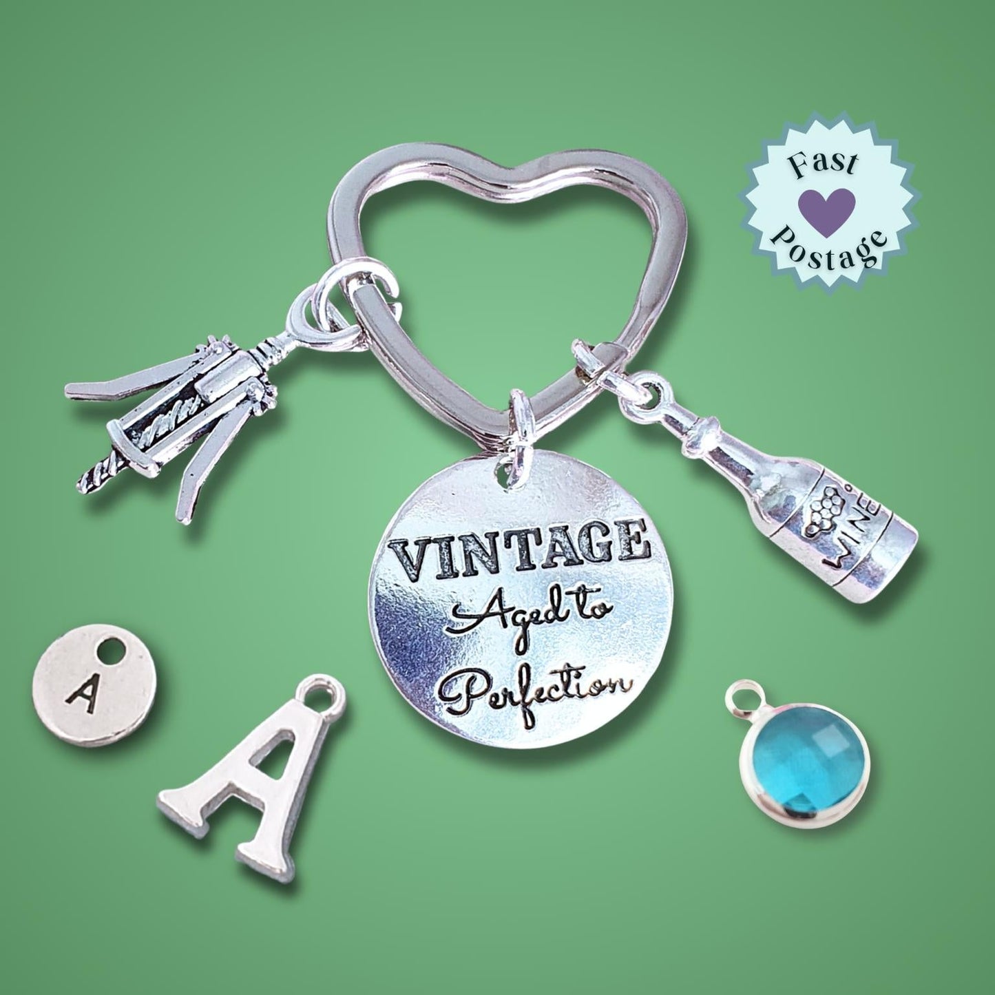 A silver-tone keychain with multiple charms, including a heart-shaped ring, a bottle charm, a champagne bottle charm, a corkscrew charm, a round engraved disc reading 'VINTAGE Aged to Perfection', a letter 'A' charm, and a blue gemstone charm, displayed on a solid green background with a 'Fast Postage' badge.