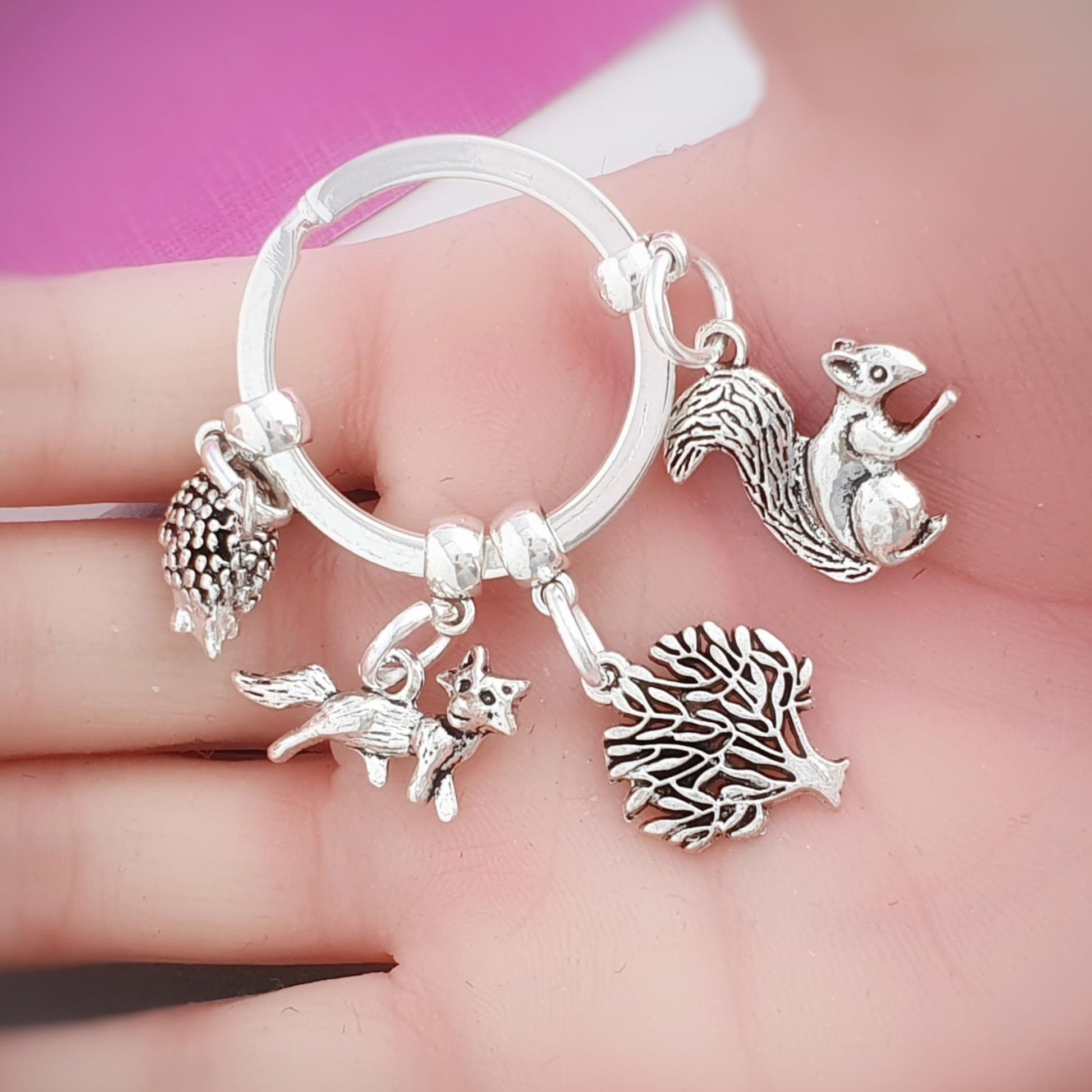 Silver keychain with squirrel, fox, tree and hedgehog charms held in a hand against a pink background