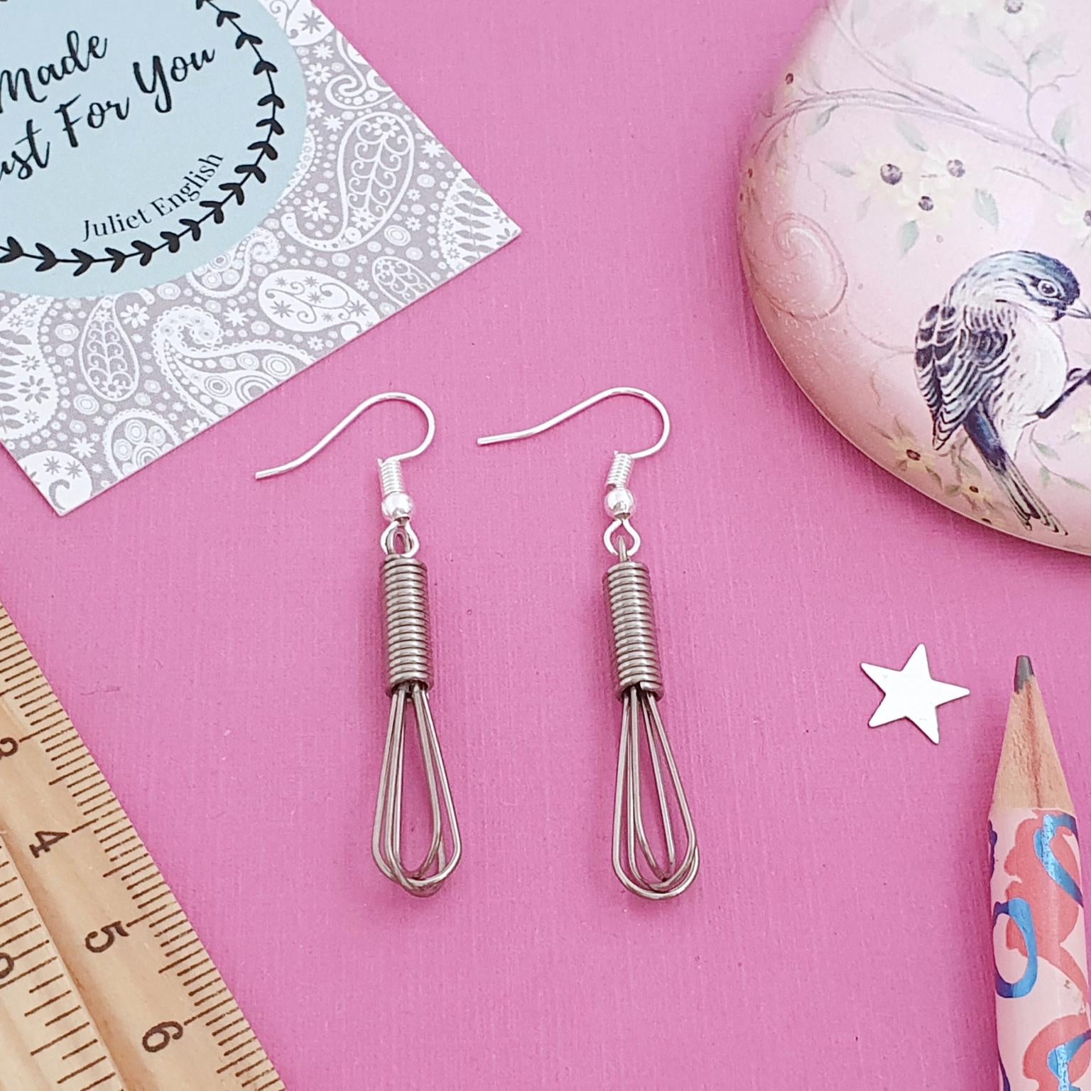 Pair of whisk-shaped earrings on a pink surface with a ruler, card, and decorative rock.
