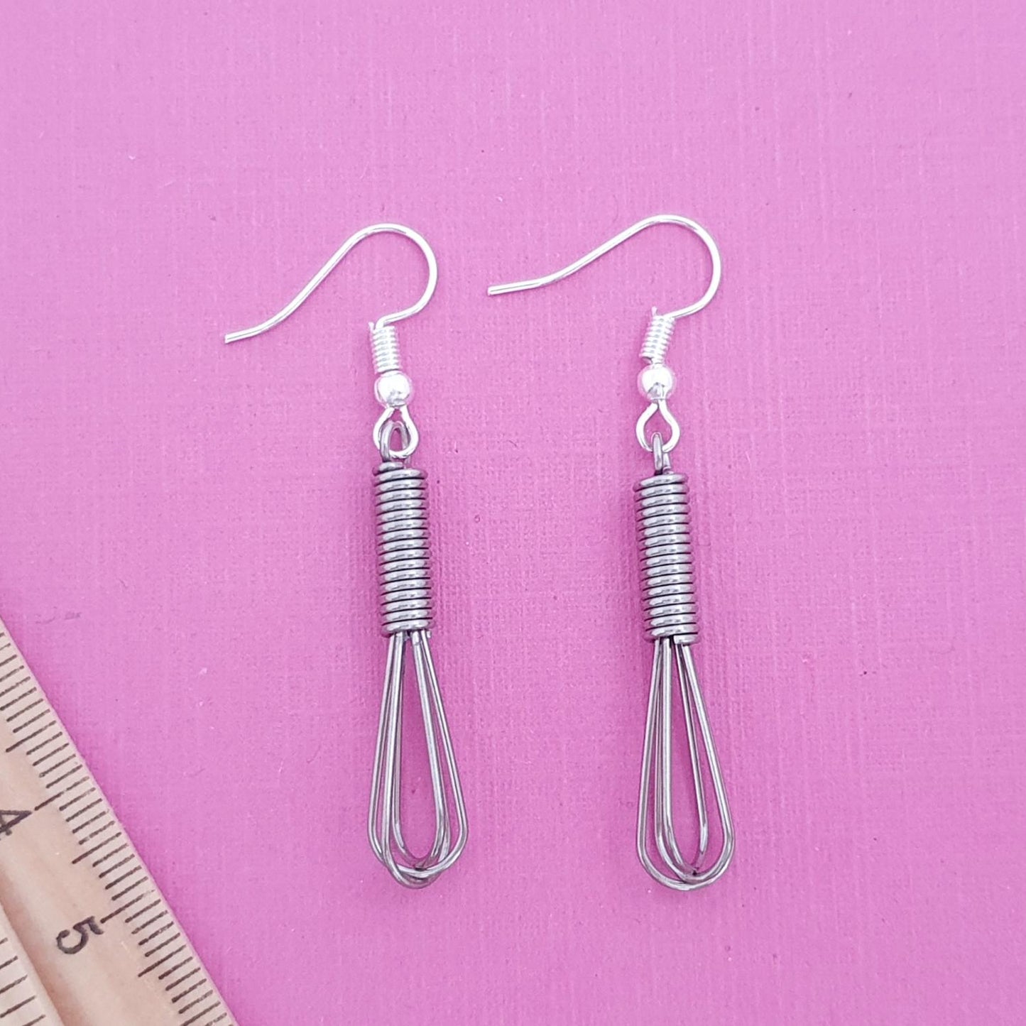 Pair of silver whisk-shaped earrings on a pink background with a ruler for scale.