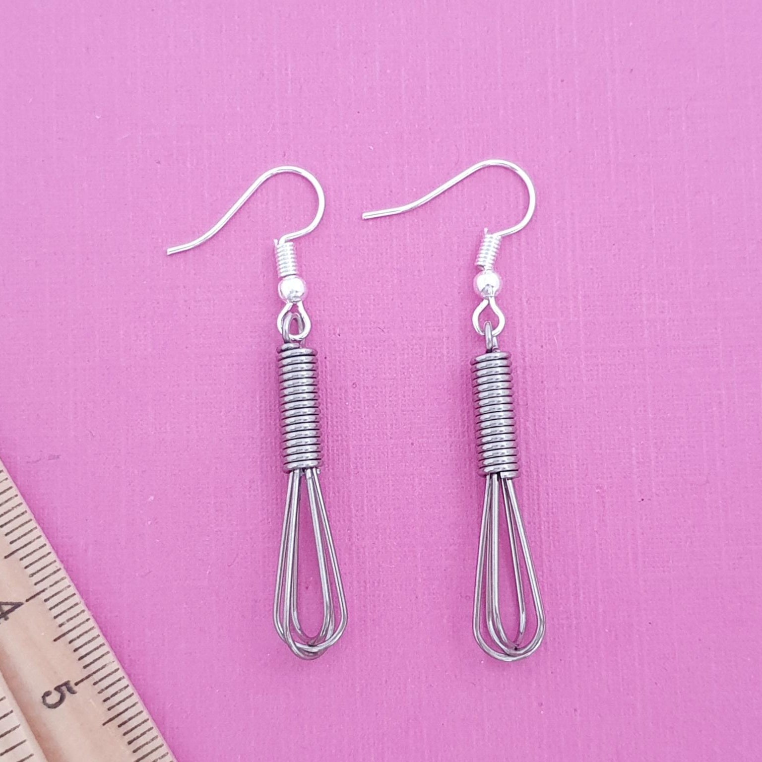 Pair of silver whisk-shaped earrings on a pink background with a ruler for scale.