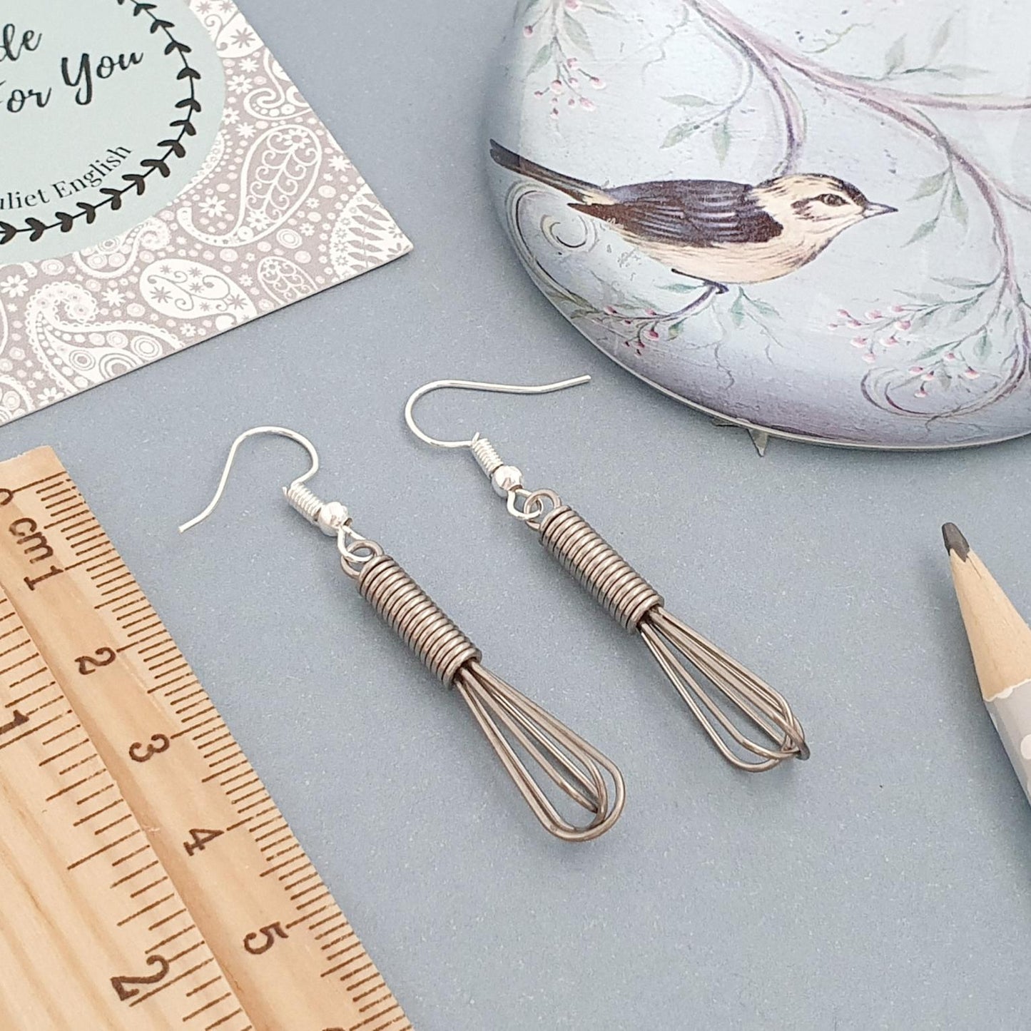 Pair of whisk earrings on a gray surface with a ruler and decorative items.