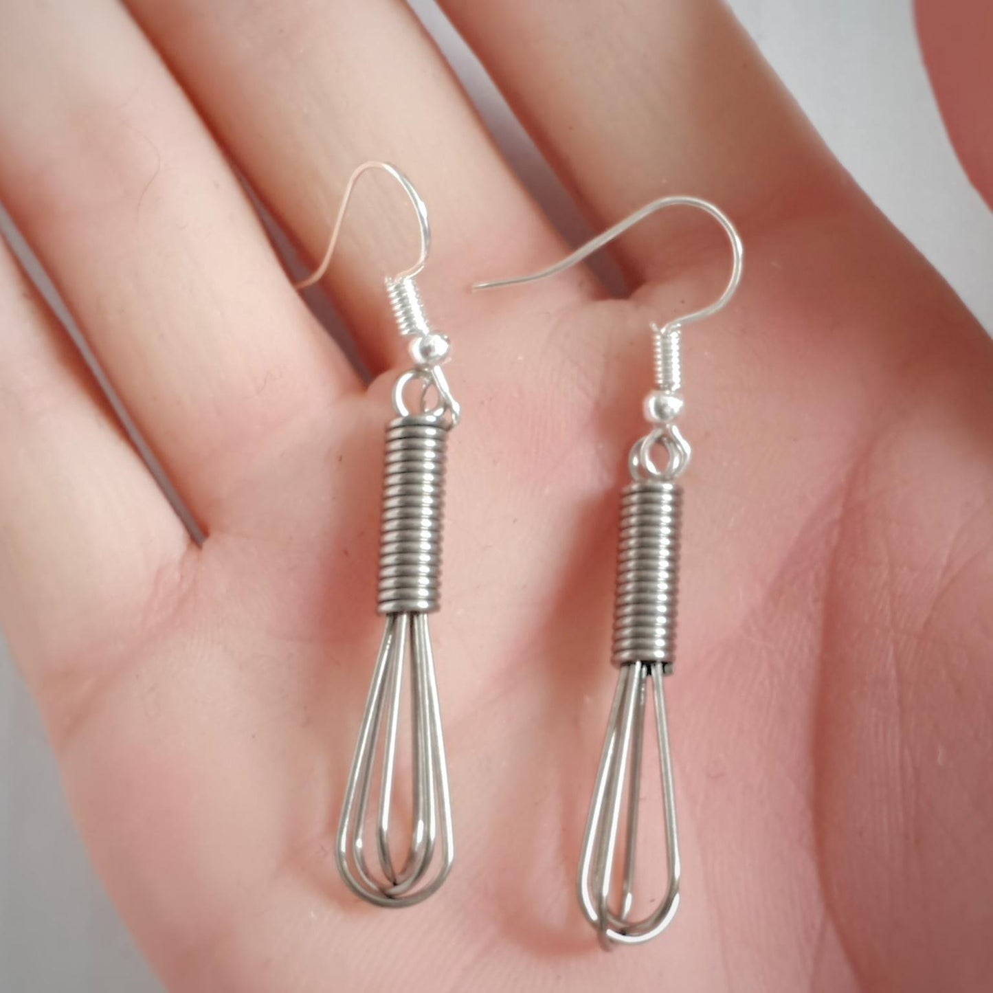 Silver whisk earrings held in a hand against a neutral background