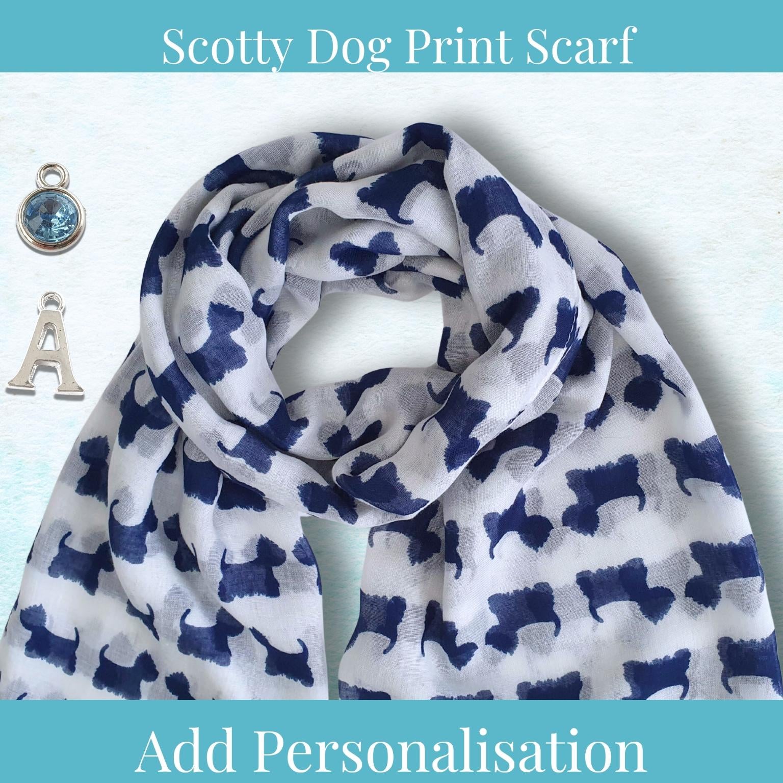 A white scarf with a navy blue Scotty Dog print pattern, styled with two silver charms (a blue gemstone pendant and a letter 'A') on a light blue textured background. Text overlays read 'Scotty Dog Print Scarf' at the top and 'Add Personalisation' at the bottom.