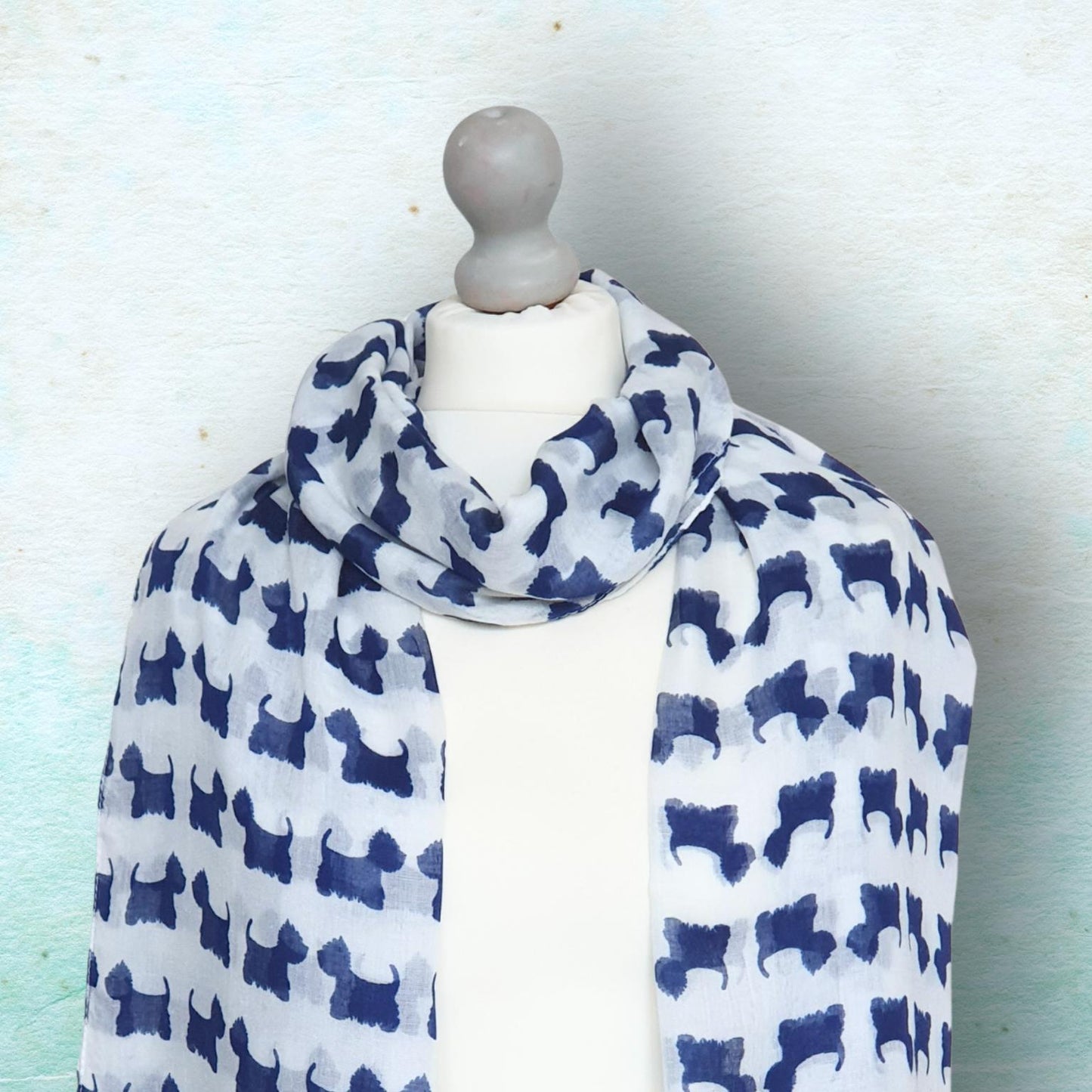 A lightweight white scarf with a repeating navy blue silhouette pattern of Scottish Terrier dogs, displayed on a white mannequin torso against a textured light blue background.