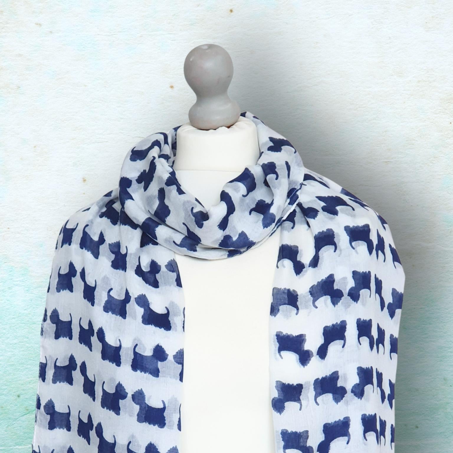 A lightweight white scarf with a repeating navy blue silhouette pattern of Scottish Terrier dogs, displayed on a white mannequin torso against a textured light blue background.