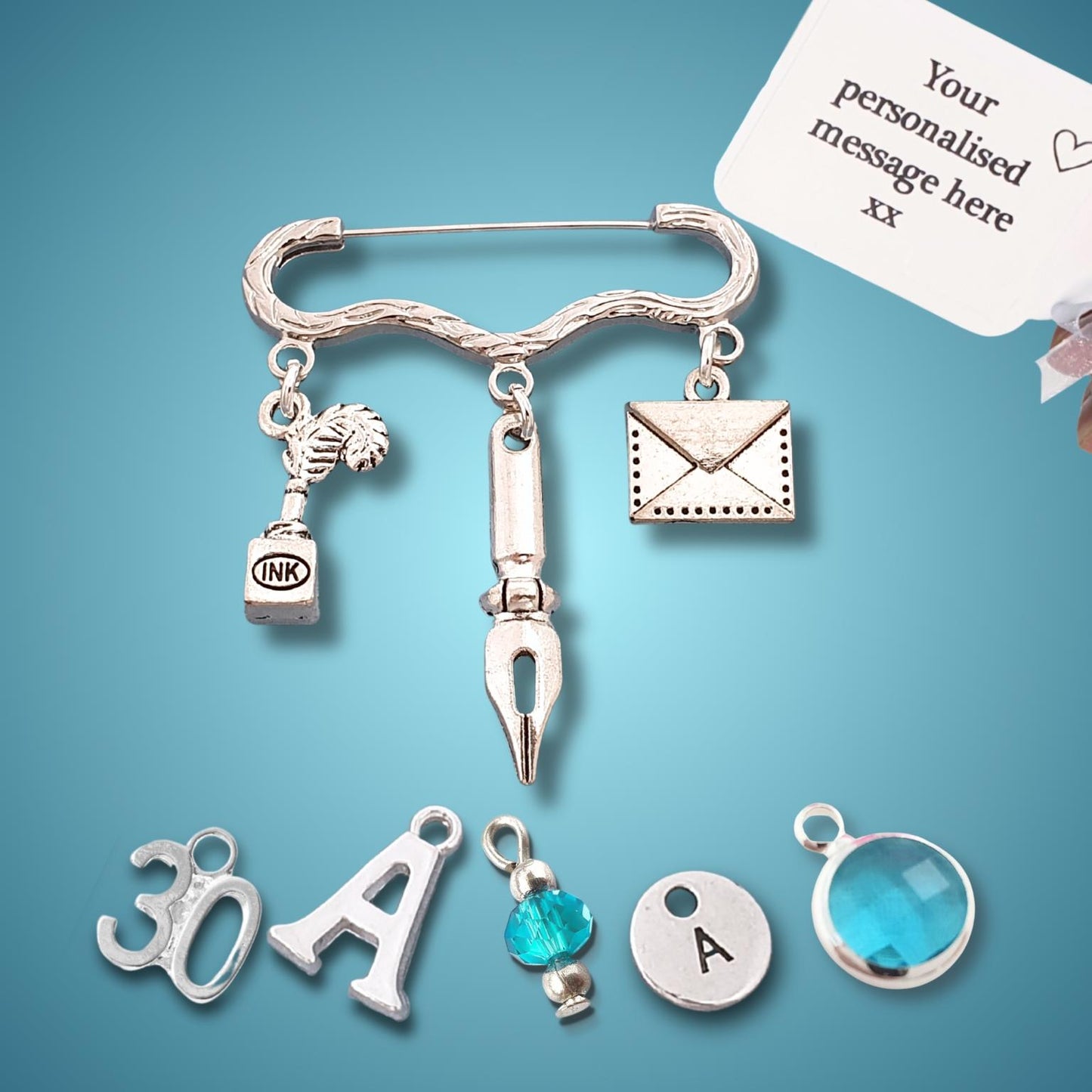 A silver personalized safety pin brooch charm set with various letter and symbol charms, shown with a hand holding a white personalization card on a blue gradient background.