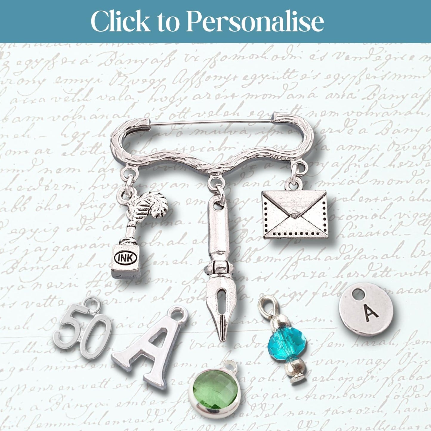 A silver-toned safety pin brooch with multiple charms, including an envelope, fountain pen nib, small bottle, green gemstone, and letter 'A' charms, displayed on a light script background with a blue 'Click to Personalise' banner at the top.