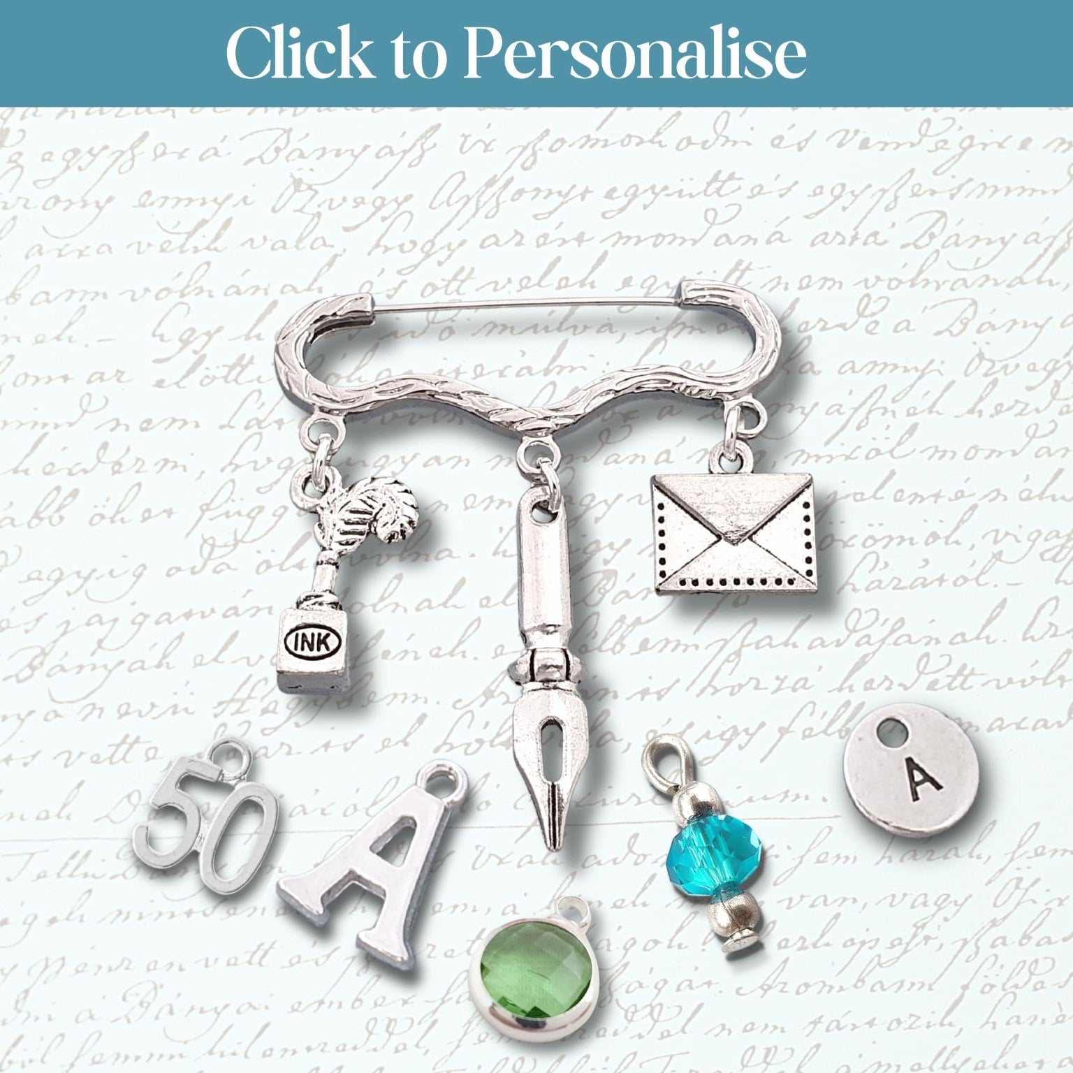 A silver-toned safety pin brooch with multiple charms, including an envelope, fountain pen nib, small bottle, green gemstone, and letter 'A' charms, displayed on a light script background with a blue 'Click to Personalise' banner at the top.