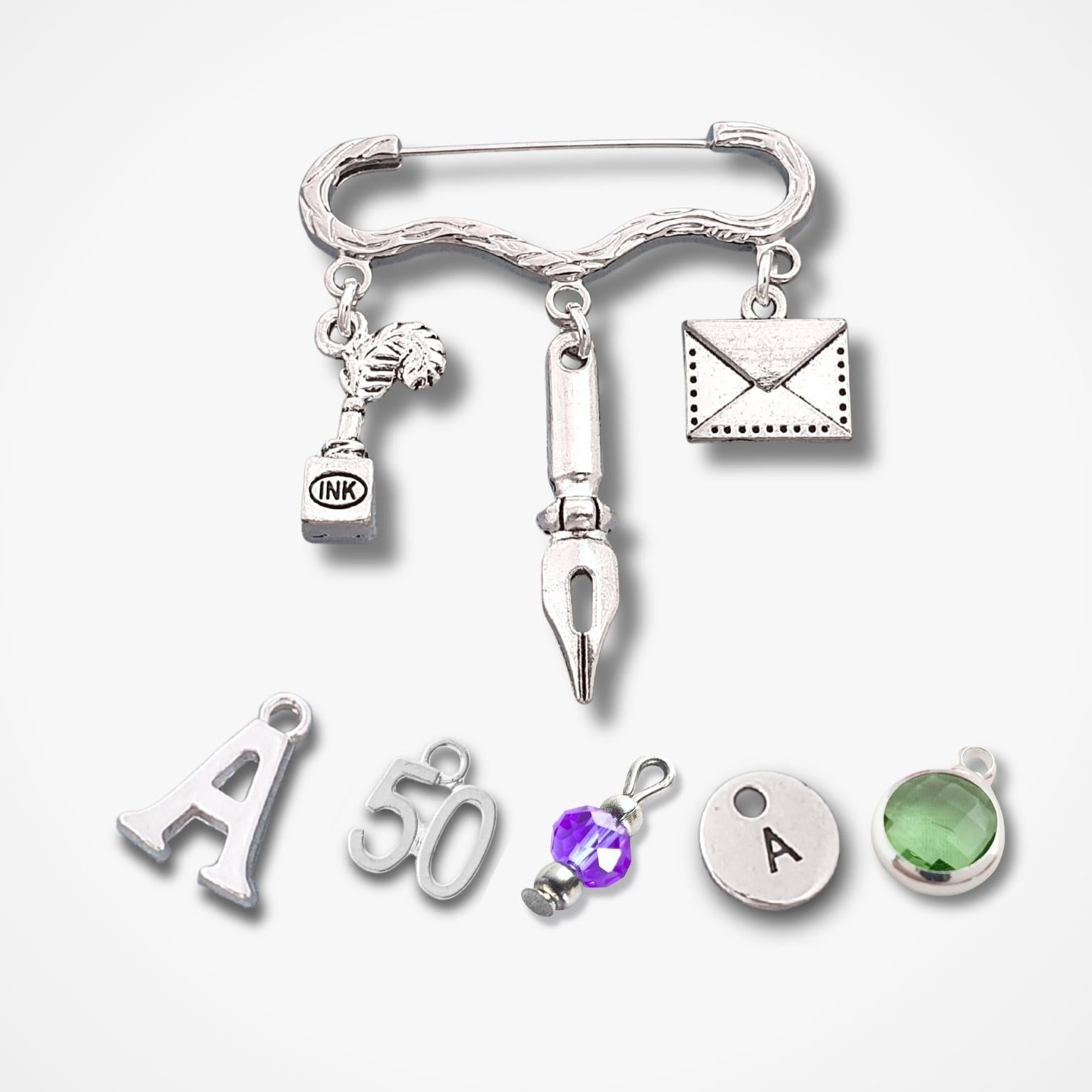 A silver-toned jewelry charm set featuring a safety pin base with dangling charms including a small trophy, a fountain pen nib, an envelope, and several loose letter and number charms, shown on a light gray background.
