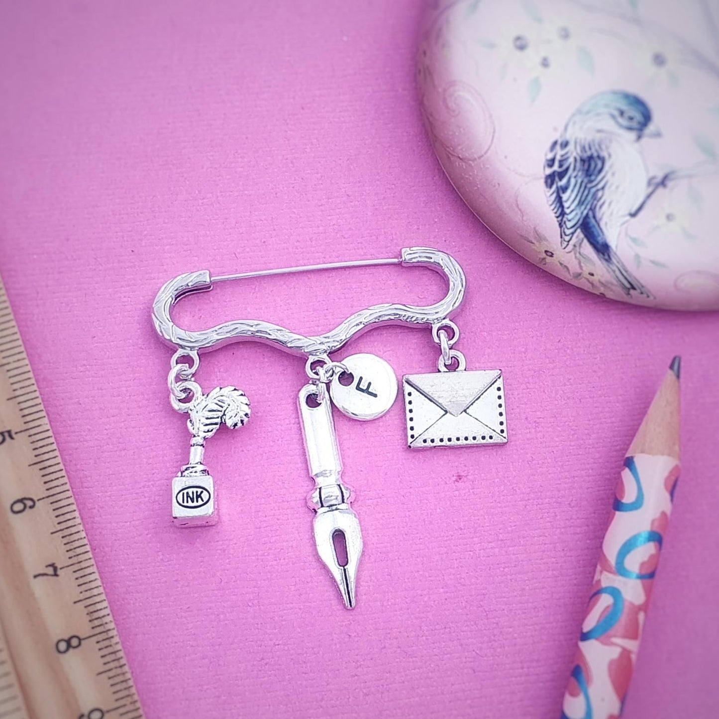 A silver-toned charm brooch shaped like a curved bar with hanging charms including a fountain pen nib, ink bottle, stamp, and envelope, displayed on a pink background with a ruler, pencil, and decorative oval object.