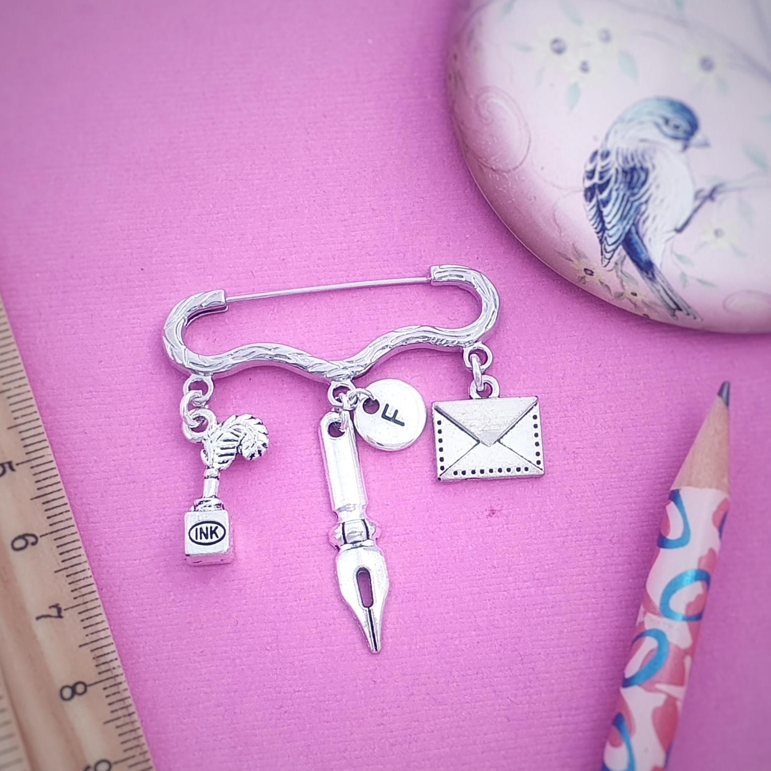 A silver-toned charm brooch shaped like a curved bar with hanging charms including a fountain pen nib, ink bottle, stamp, and envelope, displayed on a pink background with a ruler, pencil, and decorative oval object.