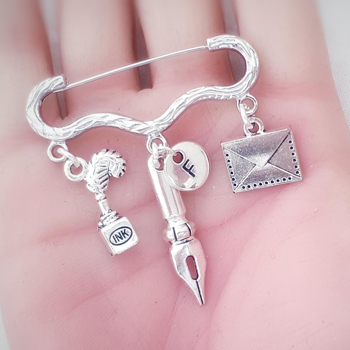 A silver-toned metal pin, held in a person's hand. The pin features dangling charms representing writing and communication: a quill with an ink bottle, a fountain pen nib, a small round charm , and an envelope.