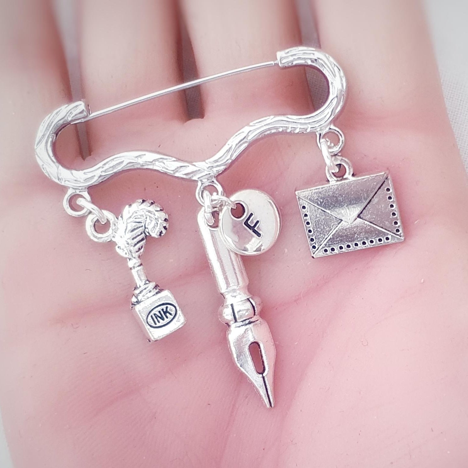 A silver-toned metal pin, held in a person's hand. The pin features dangling charms representing writing and communication: a quill with an ink bottle, a fountain pen nib, a small round charm , and an envelope.