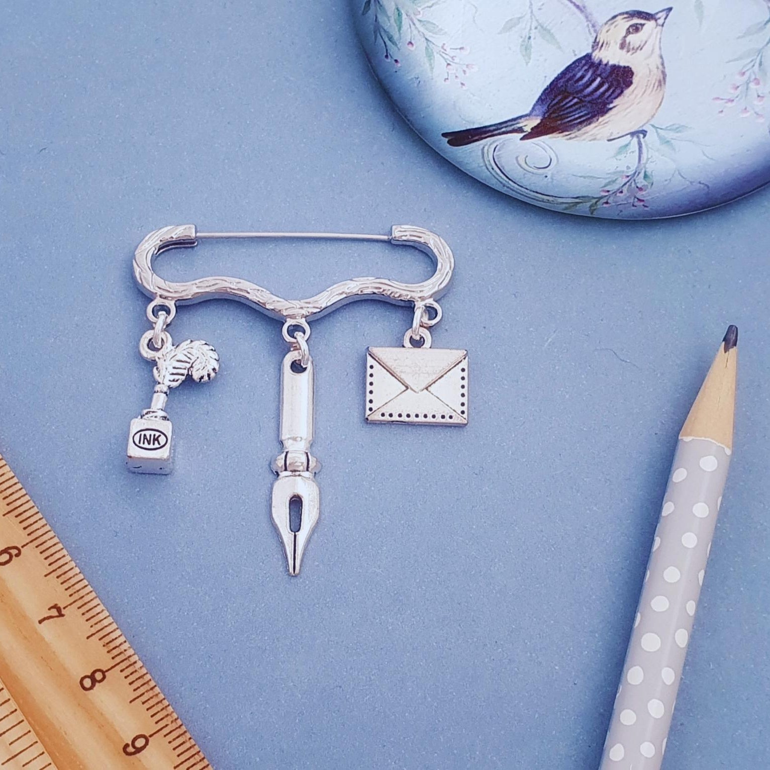 A silver-toned pin brooch with three dangling charms: a small bottle, a fountain pen nib, and an envelope, displayed on a blue background with a wooden ruler, pencil, and bird-themed round object.